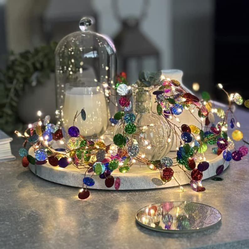 Puede incluir: Una guirnalda de adornos circulares multicolores y brillantes sobre un hilo fino, iluminada por peque&ntilde;as luces LED de color blanco c&aacute;lido. Est&aacute; dispuesta sobre una bandeja redonda de madera blanca junto a una campana de cristal, una vela y un jarr&oacute;n.