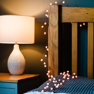 Puede incluir: Una cabecera de cama de madera con una guirnalda de luces de hadas rosas. Las luces están enhebradas en cables finos y tienen pequeñas bombillas redondas. La cama está hecha con una manta de patrón azul y blanco.