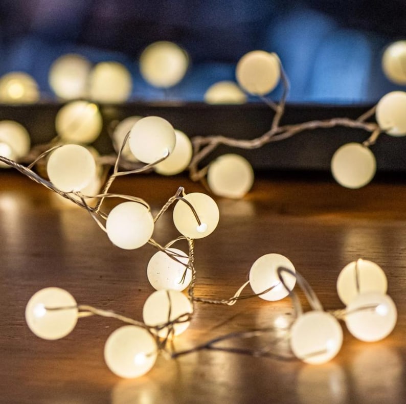 May include: String of warm white globe lights on a thin, flexible wire. The lights are evenly spaced and emit a soft glow. The string lights are arranged on a wooden surface, creating a cozy ambiance. Ideal for home decor and festive occasions.