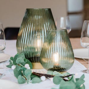 May include: Two green glass candle holders with a ribbed design. The larger candle holder has a gold base. The candle holders are on a white tablecloth with a sprig of greenery.