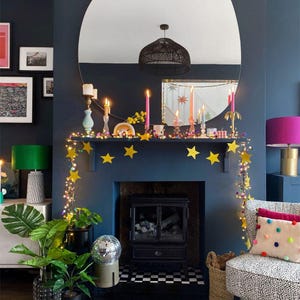 Puede incluir: Una repisa de chimenea azul oscuro decorada con una guirnalda de estrellas amarillas y luces. Varias velas c&oacute;nicas de colores en diversos candelabros se asientan sobre la repisa. Un gran espejo redondo refleja una l&aacute;mpara colgante negra.