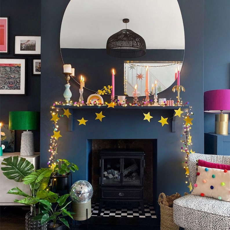 Puede incluir: Una repisa de chimenea azul oscuro decorada con una guirnalda de estrellas amarillas y luces. Varias velas c&oacute;nicas de colores en diversos candelabros se asientan sobre la repisa. Un gran espejo redondo refleja una l&aacute;mpara colgante negra.