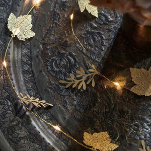 May include: A string of warm white fairy lights with gold leaf cutouts of leaves and branches. The lights are strung on a dark background.