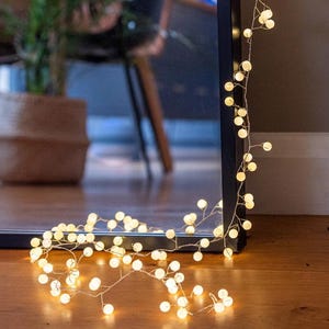May include: A string of warm white LED globe lights, draped along a dark frame and wooden floor. The lights are arranged in a delicate, vine-like pattern, creating a soft, ambient glow. The lights are reflected in a dark mirror.