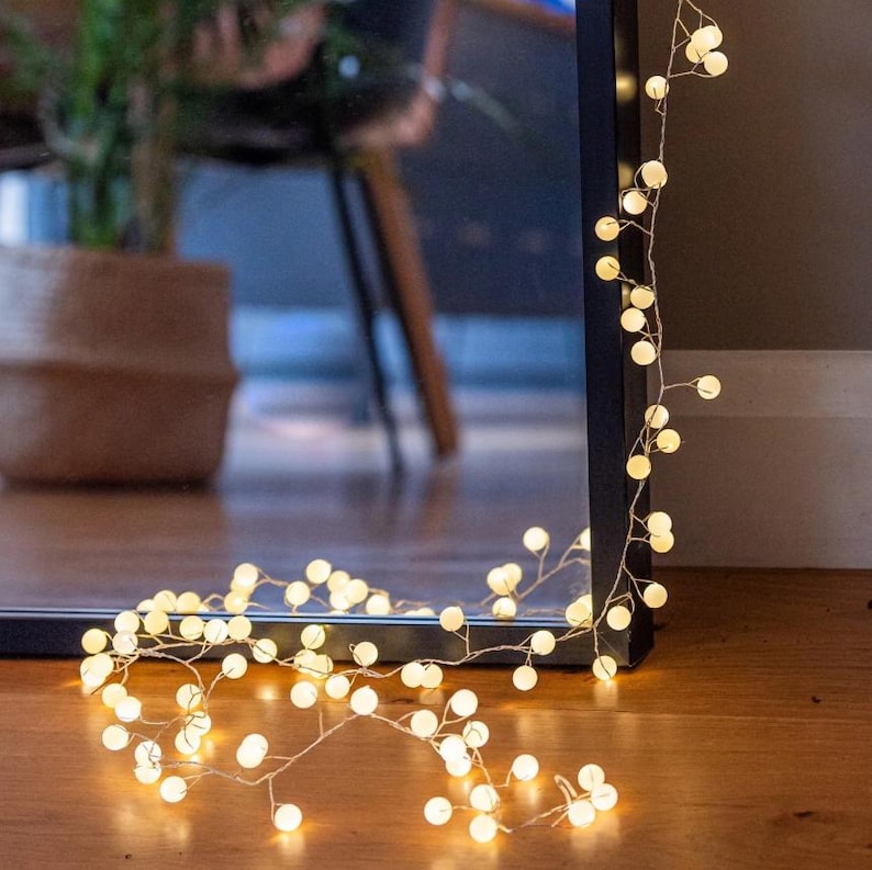 May include: A string of warm white LED globe lights, draped along a dark frame and wooden floor. The lights are arranged in a delicate, vine-like pattern, creating a soft, ambient glow. The lights are reflected in a dark mirror.