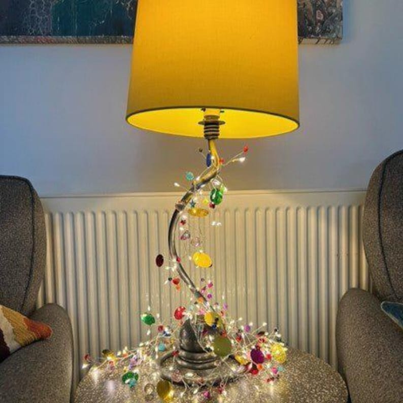 Puede incluir: Una l&aacute;mpara de mesa con una pantalla cil&iacute;ndrica de color amarillo s&oacute;lido y una base plateada curva. La base est&aacute; envuelta en finos hilos de alambre con peque&ntilde;as cuentas transl&uacute;cidas multicolores y diminutas luces LED integradas.