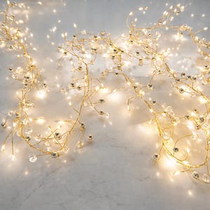 Peut inclure: Une guirlande lumineuse décorative composée d'un fil fin et flexible de couleur dorée, entrelacé de petites perles sphériques transparentes et de lumières LED blanc chaud qui diffusent un éclairage doux sur toute la longueur du cordon.