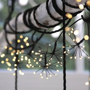 Pode incluir: Um fio de luzes decorativas com fios pretos finos e pequenas lâmpadas LED de cor branca quente dispostas em grupos esféricos semelhantes a fogo de artifício. As luzes estão enroladas em torno de um ramo ou poste de madeira de cor clara.