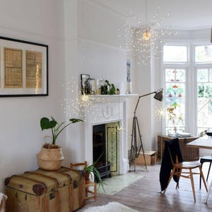 May include: A bright living room featuring starburst lights, a white fireplace, and a vintage trunk. A framed picture hangs on the wall. A floor lamp and a wooden chair add to the room's decor. A potted plant sits on the trunk. The room has wooden flooring.