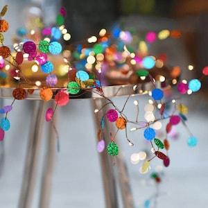 May include: A string of fairy lights with colourful, iridescent, round ornaments. The lights are strung on a copper wire and are hanging from a table.