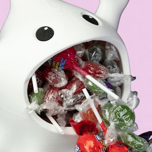 May include: A white, cartoon-style candy dispenser with black eyes and a wide mouth filled with wrapped lollipops. The lollipops are in various colours, including red, green, and purple, with white sticks.