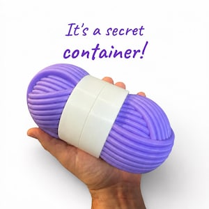May include: A purple, yarn-shaped container with a smooth white band around the centre. The text "It's a secret container!" is printed in purple above the object. The container features a textured, coiled design resembling a ball of yarn.