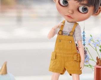 Puede incluir: Muñeca con cabello gris y ojos azules grandes, vestida con un conjunto de peto amarillo, camiseta blanca y zapatillas negras y blancas. Una pequeña estantería exhibe libros y un conejito de ganchillo azul. Un letrero dice "Ice Cream".