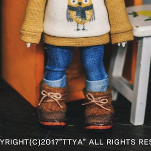 May include: A doll with brown curly hair wearing a yellow and white sweater with an owl design and blue jeans. The doll is wearing brown boots and has pink hair clips. The doll is standing in front of a red wooden cabinet with the word "ACKNOWL" written on it.