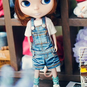 May include: A doll with red hair wearing a blue beanie, a white shirt, and denim overalls. The doll is standing on a wooden floor with a small chair and a bag in the background.