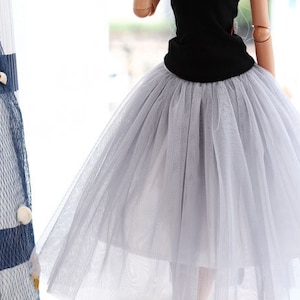 May include: A doll wearing a black tank top and a light gray tulle skirt. The doll has blonde hair and blue eyes. The doll is wearing blue and white striped socks and blue sneakers with white laces. The doll is standing in front of a window.
