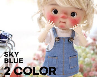 May include: A doll with blonde hair and rosy cheeks wearing a white t-shirt under blue denim overalls with two front pockets and brown buttons. The text SKY BLUE 2 COLOR is visible on the left side of the image near a red and white toy vehicle.