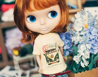 Puede incluir: Una muñeca con cabello castaño rojizo hasta los hombros y ojos azul brillante, que viste una camiseta color crema con un gráfico de una furgoneta vintage, jeans azules con un cinturón rojo fino, leggings a rayas verdes y blancas y botas marrones.