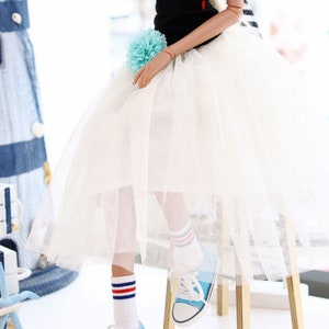 K&ouml;nnte beinhalten: Eine Puppe tr&auml;gt ein schwarzes Tanktop, einen wei&szlig;en T&uuml;llrock, blau-wei&szlig; gestreifte Socken und blaue Turnschuhe. Die Puppe hat blondes Haar und sitzt auf einem Holzhocker. Die Puppe steht vor einem Fenster mit einem blau-wei&szlig; gestreiften Vorhang.