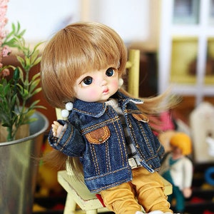 以下が含まれることがあります： 青いデニムジャケット、黄色のズボン、白いスニーカーを履いた小さな人形が木製の椅子に座っています。人形はブロンドの髪と青い目をしています。椅子は黒と黄色の模様の敷物の上にあります。