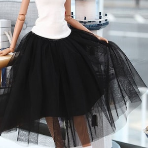 May include: A doll wearing a white tank top and a black tulle skirt. The doll has long pink hair and is wearing blue and white striped socks and blue sneakers. The doll is sitting on a wooden chair.