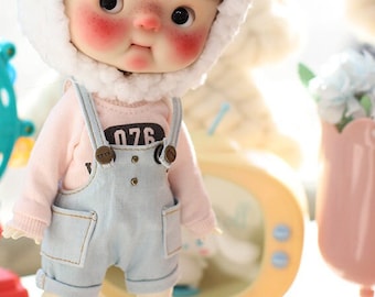 May include: A small doll wearing a white and black panda hat, a pink long-sleeved shirt, blue overalls, and white sneakers. The doll has brown hair and rosy cheeks.