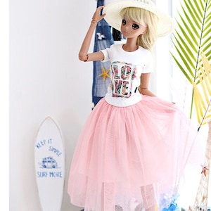 May include: A doll wearing a white t-shirt with a floral "LOVE" graphic, a pink tulle skirt, yellow sneakers, and white and red striped socks. The doll is wearing a white straw hat and has blonde hair. The doll is standing in front of a white background with a lighthouse and palm tree.