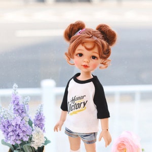 Puede incluir: Una mu&ntilde;eca con el pelo rojo en coletas que lleva una camiseta de b&eacute;isbol blanca y negra con las palabras "Honor Victory" impresas. La mu&ntilde;eca lleva pantalones cortos de mezclilla azul, calcetines blancos y zapatos marrones. La mu&ntilde;eca est&aacute; de pie sobre una superficie blanca con un oso de peluche, una manzana y dos juguetes amarillos de pollitos.