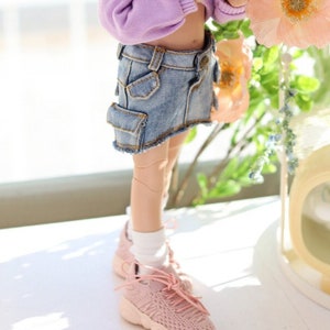 Pode incluir: Uma boneca rosa com cabelo rosa em tran&ccedil;as usando um moletom roxo com capuz e uma saia jeans azul. A boneca est&aacute; segurando uma flor rosa e est&aacute; em p&eacute; em uma superf&iacute;cie branca. A boneca est&aacute; usando t&ecirc;nis rosa. COPYRIGHT (C) 2022 "TTYA" ALL RIGHTS RESERVED