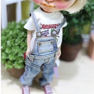 May include: A doll with blonde hair and rosy cheeks wearing a white t-shirt with "AVENGERS CAPTAIN AMERICA" text, blue denim overalls, and pink sneakers. The doll is standing in front of a green plant.