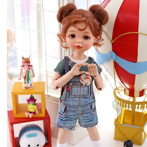 May include: A doll with red hair in pigtails wearing a blue denim overall dress and a green and white patterned shirt. The doll is holding a colorful lollipop and is standing in front of a red and white hot air balloon.