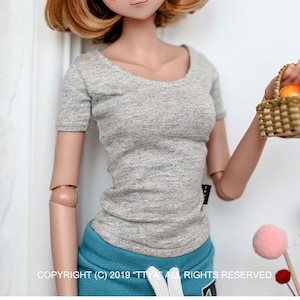 May include: A doll with light brown hair and large, expressive eyes, wearing a gray short-sleeved shirt and blue shorts. A red patterned headband adorns the doll's head. The doll holds a small basket with fruit.