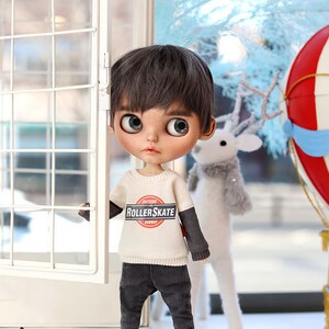 May include: A doll wearing a cream colored sweater with the words "Outside the Roller Skate Runway" printed on the front. The doll is wearing black pants and black sneakers. The doll has brown hair and blue eyes.