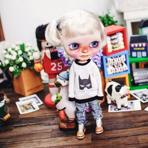 May include: A white doll with blonde hair in pigtails wearing a white sweater with a black bat graphic and blue jeans. The doll is standing next to a white goose figurine and a brown and white cow figurine.