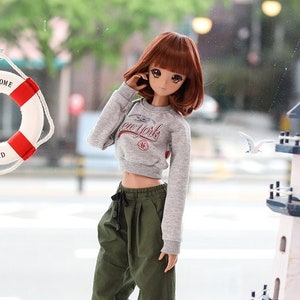 May include: A doll wearing a grey cropped sweatshirt with the text "New York" on the front, green cargo pants, and white sneakers. The doll is standing in front of a window with a life preserver and a miniature lighthouse.