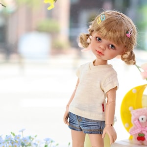 May include: A doll with blonde hair wearing a white t-shirt, blue denim shorts, and pink and blue sneakers. The doll is standing on a white surface with a small wooden bench behind it. The bench has a pink teddy bear and a sign that says "Ice Cream Cold Treat".