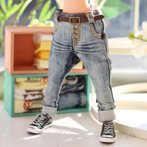 May include: A doll wearing a pair of light blue denim jeans with a brown belt and black sneakers. The doll is standing in front of a shelf with stacked clothing.