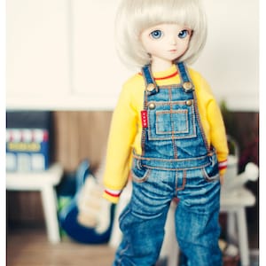 May include: A doll with a platinum blonde bob haircut and blue eyes, wearing a yellow long-sleeved shirt and denim overalls. The doll is also wearing yellow shoes. The overalls have metal clasps and pockets.