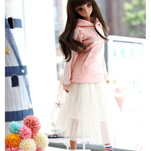 K&ouml;nnte beinhalten: Eine Puppe tr&auml;gt ein rosa Hoodie mit wei&szlig;en Streifen, einen wei&szlig;en T&uuml;llrock, wei&szlig;e Socken mit roten und blauen Streifen und blaue Turnschuhe. Die Puppe hat langes braunes Haar und steht vor einem Fenster.