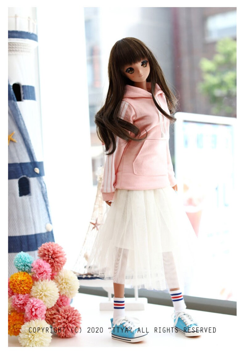 K&ouml;nnte beinhalten: Eine Puppe tr&auml;gt ein rosa Hoodie mit wei&szlig;en Streifen, einen wei&szlig;en T&uuml;llrock, wei&szlig;e Socken mit roten und blauen Streifen und blaue Turnschuhe. Die Puppe hat langes braunes Haar und steht vor einem Fenster.