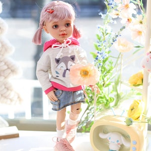 Pode incluir: Uma boneca de cabelo rosa usando um moletom cinza com um gr&aacute;fico de raposa, detalhes rosa e uma saia jeans. A boneca est&aacute; segurando uma flor rosa e est&aacute; em p&eacute; em uma superf&iacute;cie branca. H&aacute; uma televis&atilde;o amarela com um personagem de desenho animado branco dentro.