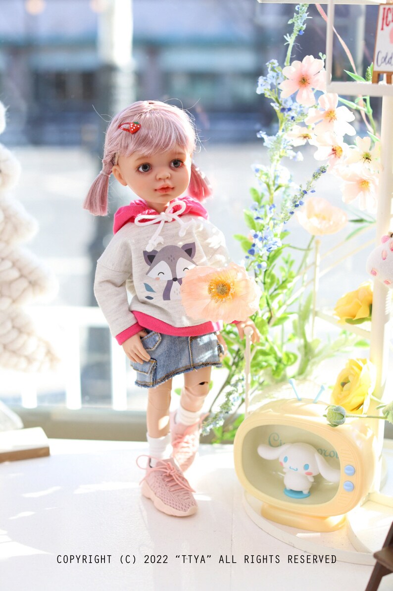 Pode incluir: Uma boneca de cabelo rosa usando um moletom cinza com um gr&aacute;fico de raposa, detalhes rosa e uma saia jeans. A boneca est&aacute; segurando uma flor rosa e est&aacute; em p&eacute; em uma superf&iacute;cie branca. H&aacute; uma televis&atilde;o amarela com um personagem de desenho animado branco dentro.