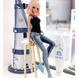 Puede incluir: Una mu&ntilde;eca con un top negro sin mangas, jeans azules y zapatillas azules est&aacute; sentada en un taburete de madera. La mu&ntilde;eca tiene el pelo largo y rosa y lleva una diadema amarilla. En el fondo hay una tabla de surf blanca con el texto "Keep it simple, surf more".