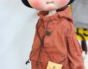 Puede incluir: Una muñeca con mejillas rosadas y un sombrero negro lleva una chaqueta color óxido con capucha y un pequeño bolso amarillo claro. La chaqueta tiene cierre de botón y cordones grises oscuros. Los ojos de la muñeca son grandes y negros.