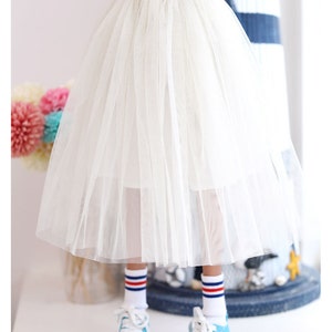 K&ouml;nnte beinhalten: Ein wei&szlig;er T&uuml;llrock mit einem goldenen Glitzerbund. Der Rock ist geschichtet und hat eine flie&szlig;ende Silhouette. Der Rock wird von einer Puppe getragen.