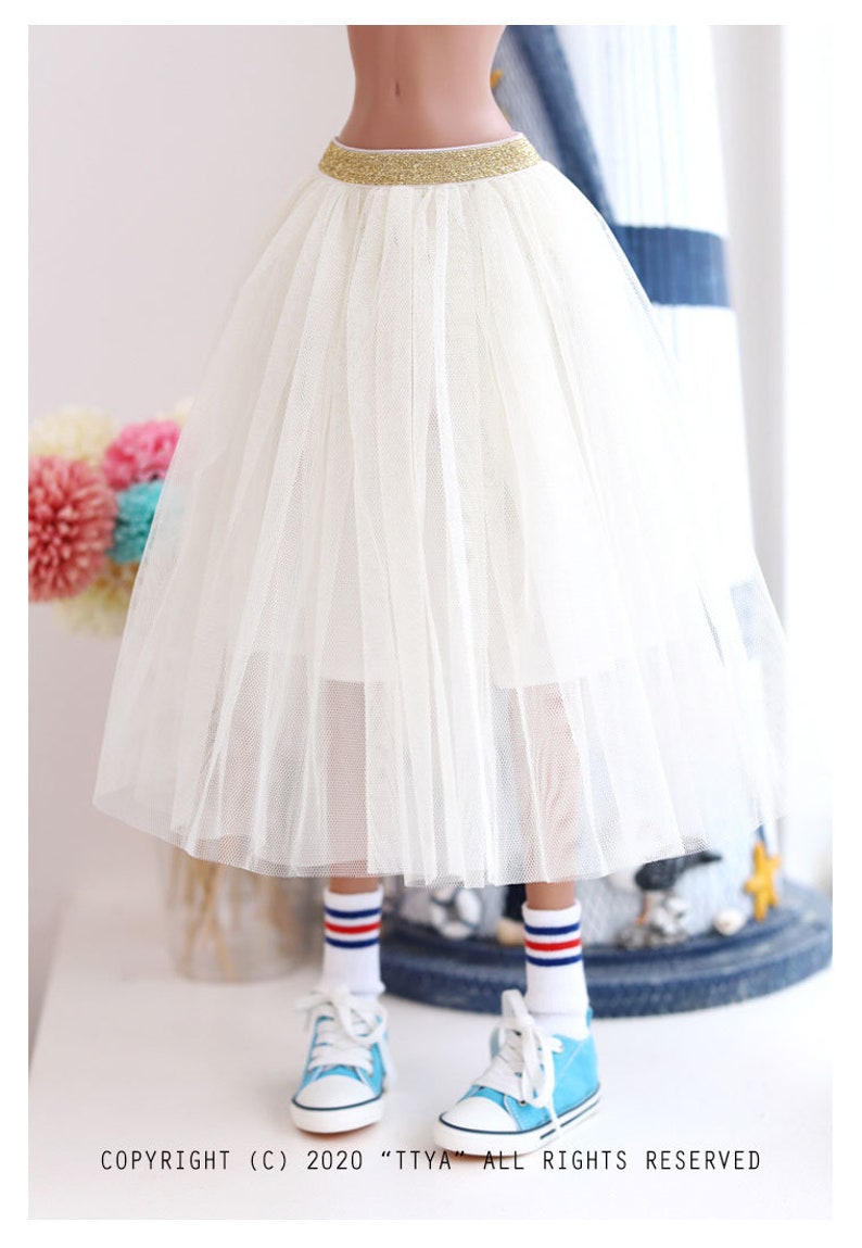 K&ouml;nnte beinhalten: Ein wei&szlig;er T&uuml;llrock mit einem goldenen Glitzerbund. Der Rock ist geschichtet und hat eine flie&szlig;ende Silhouette. Der Rock wird von einer Puppe getragen.