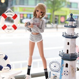 May include: A doll wearing a grey sweater with the words "New York" on it, grey shorts, black and white striped socks, and white sneakers. The doll is holding a camera and is standing in front of a white lighthouse with blue accents.