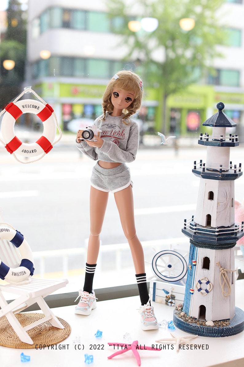 May include: A doll wearing a grey sweater with the words "New York" on it, grey shorts, black and white striped socks, and white sneakers. The doll is holding a camera and is standing in front of a white lighthouse with blue accents.