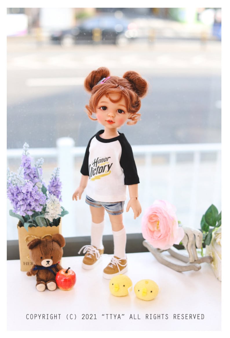 Puede incluir: Una mu&ntilde;eca con el pelo rojo en coletas que lleva una camiseta de b&eacute;isbol blanca y negra con las palabras "Honor Victory" impresas. La mu&ntilde;eca lleva pantalones cortos de mezclilla azul, calcetines blancos y zapatos marrones. La mu&ntilde;eca est&aacute; de pie sobre una superficie blanca con un oso de peluche, una manzana y dos juguetes amarillos de pollitos.