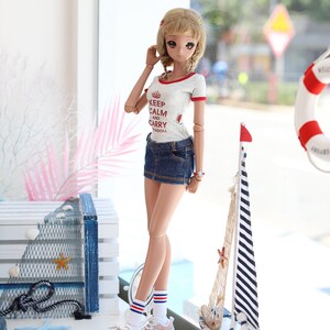 Puede incluir: Una mu&ntilde;eca con una camiseta blanca con texto rojo que dice "Keep Calm and Carry Yadoll", pantalones cortos de mezclilla azul, calcetines blancos con rayas rojas y azules y zapatillas de deporte rosa y blancas. La mu&ntilde;eca est&aacute; de pie frente a una ventana con un velero y un salvavidas visibles en el fondo.